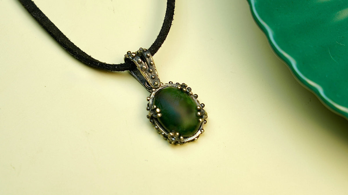A deep green jade oval on a black cord lying on pale yellow, the polished stone ringed by small silver beads