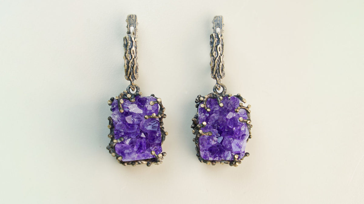 A pair of amethyst earrings with square raw crystal faces, hanging side by side on a cream background