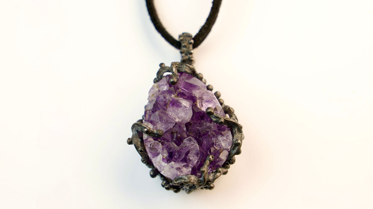 An amethyst cluster held in a dark silver frame on a black cord, the points violet at the tips and pale underneath