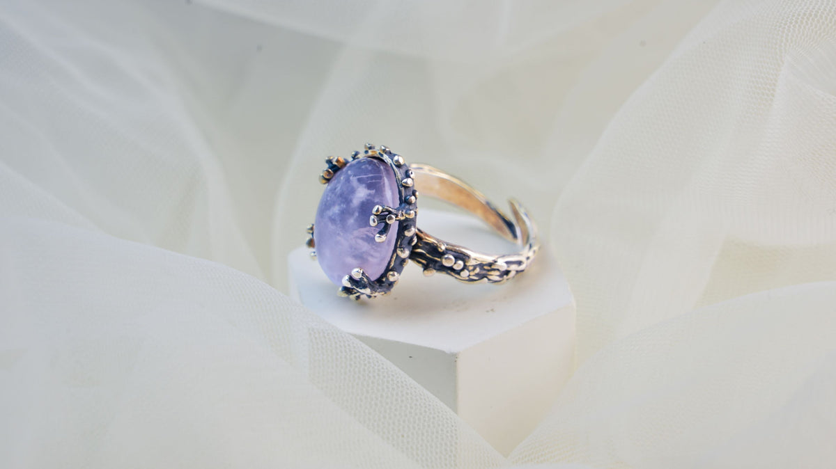 A rose quartz oval on a white block over soft tulle, a cloud drifting through the polished lilac dome