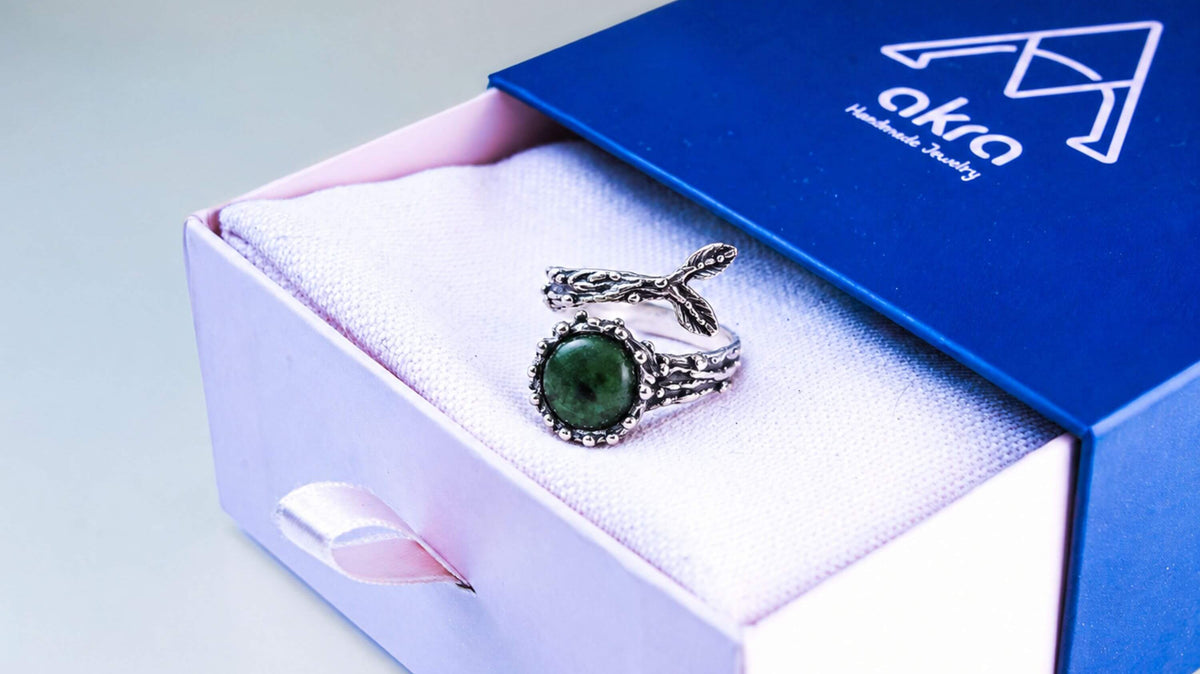 A sterling silver ring with a round green jade and a two-leaf sprig, resting on the cushion of an open blue Akra gift box.