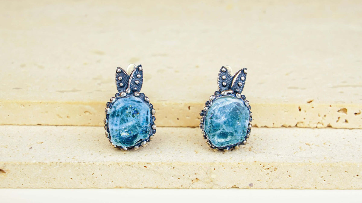 Two pale blue aquamarine cushions standing on a stone ledge, each in a beaded rim below a small silver leaf