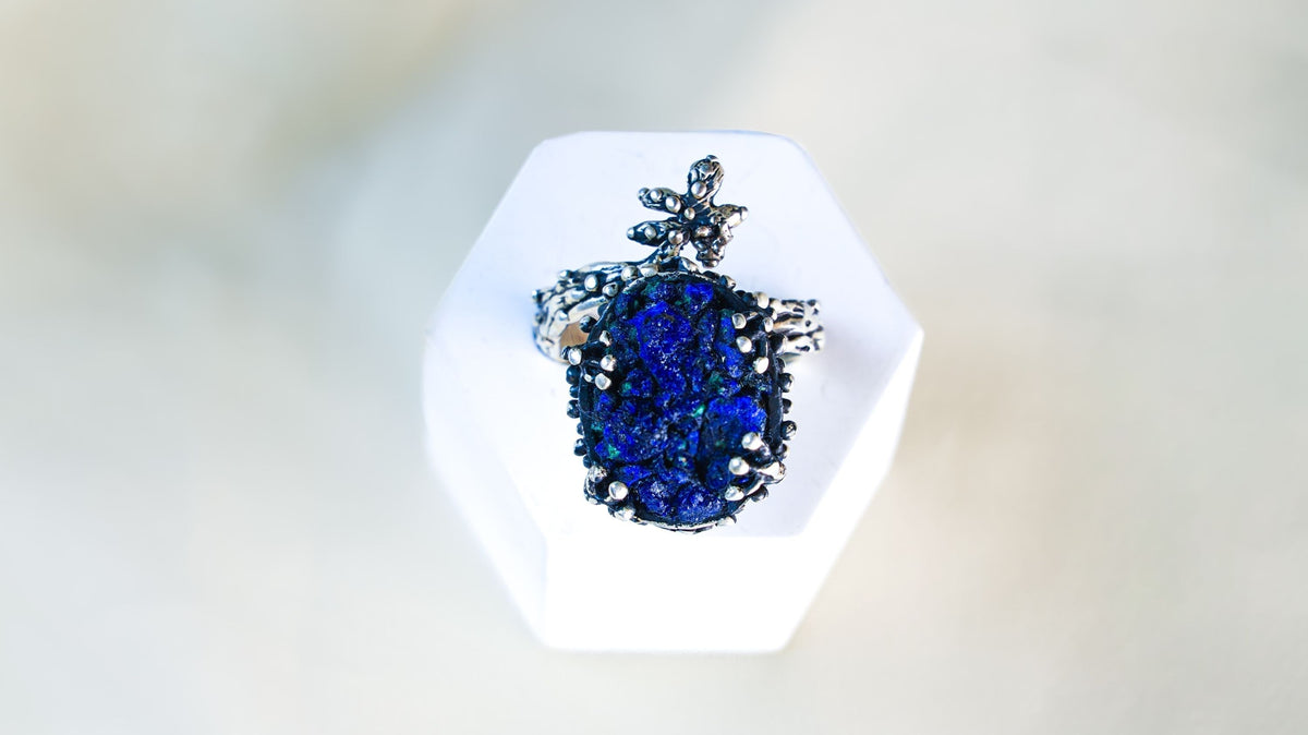 A rough blue lapis lazuli standing on a white hexagon block, the silver sprig curving above its uneven crystal face