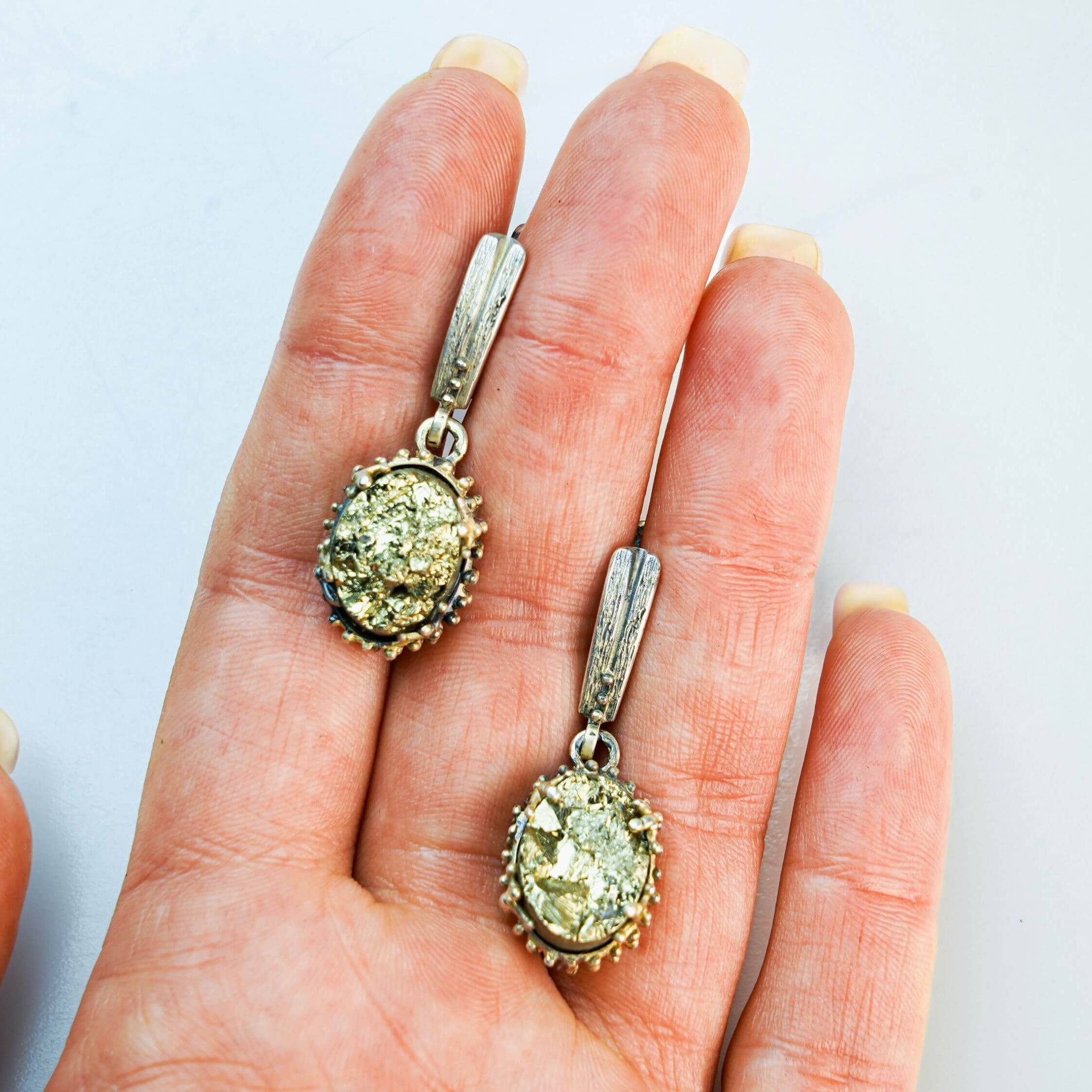 Two golden pyrite statement earrings held in a hand to illustrate their perfect oval scale and handcrafted silver detailing