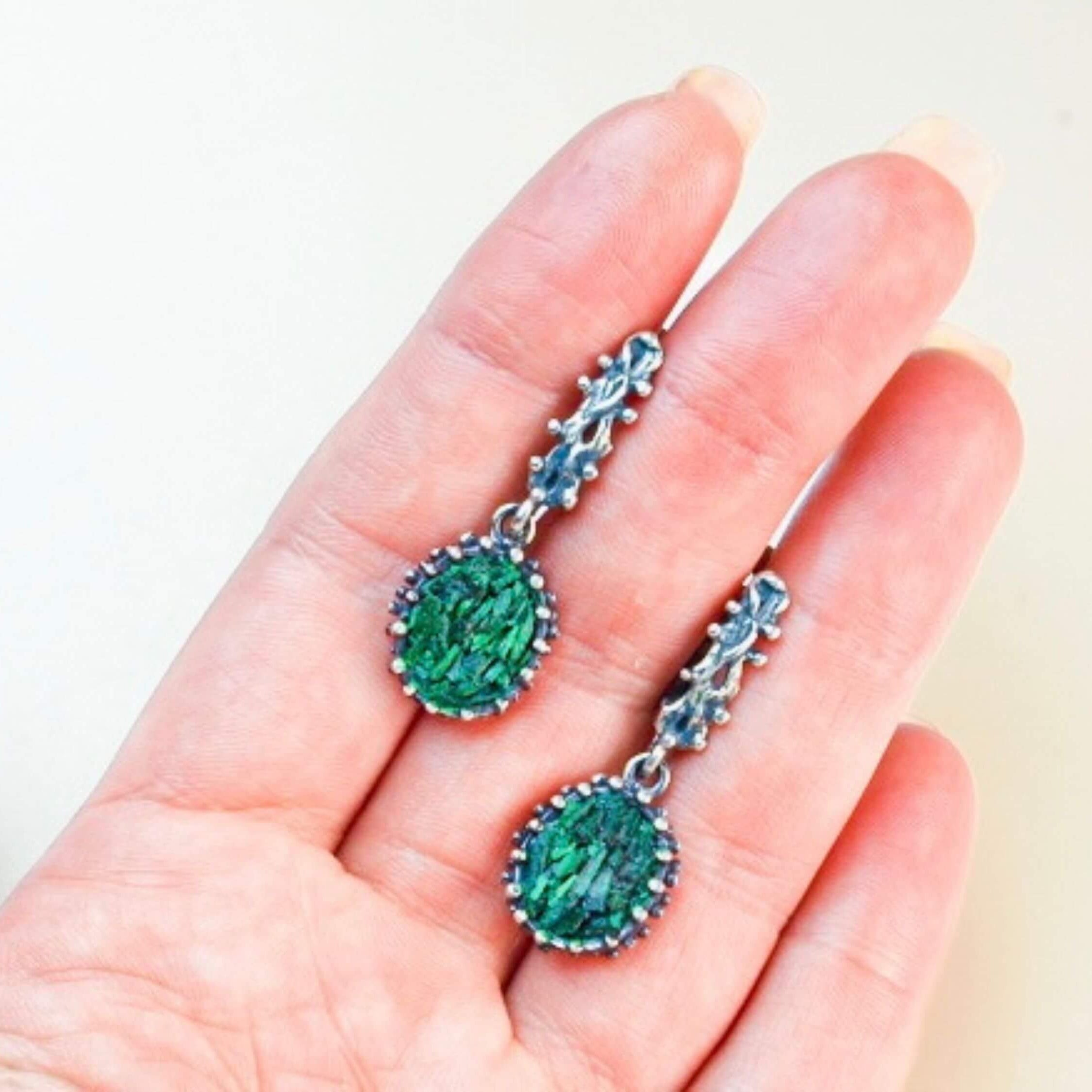 A pair of Akra Gems handcrafted oval Malachite earrings held in a hand to demonstrate the physical scale and artisan craftsmanship