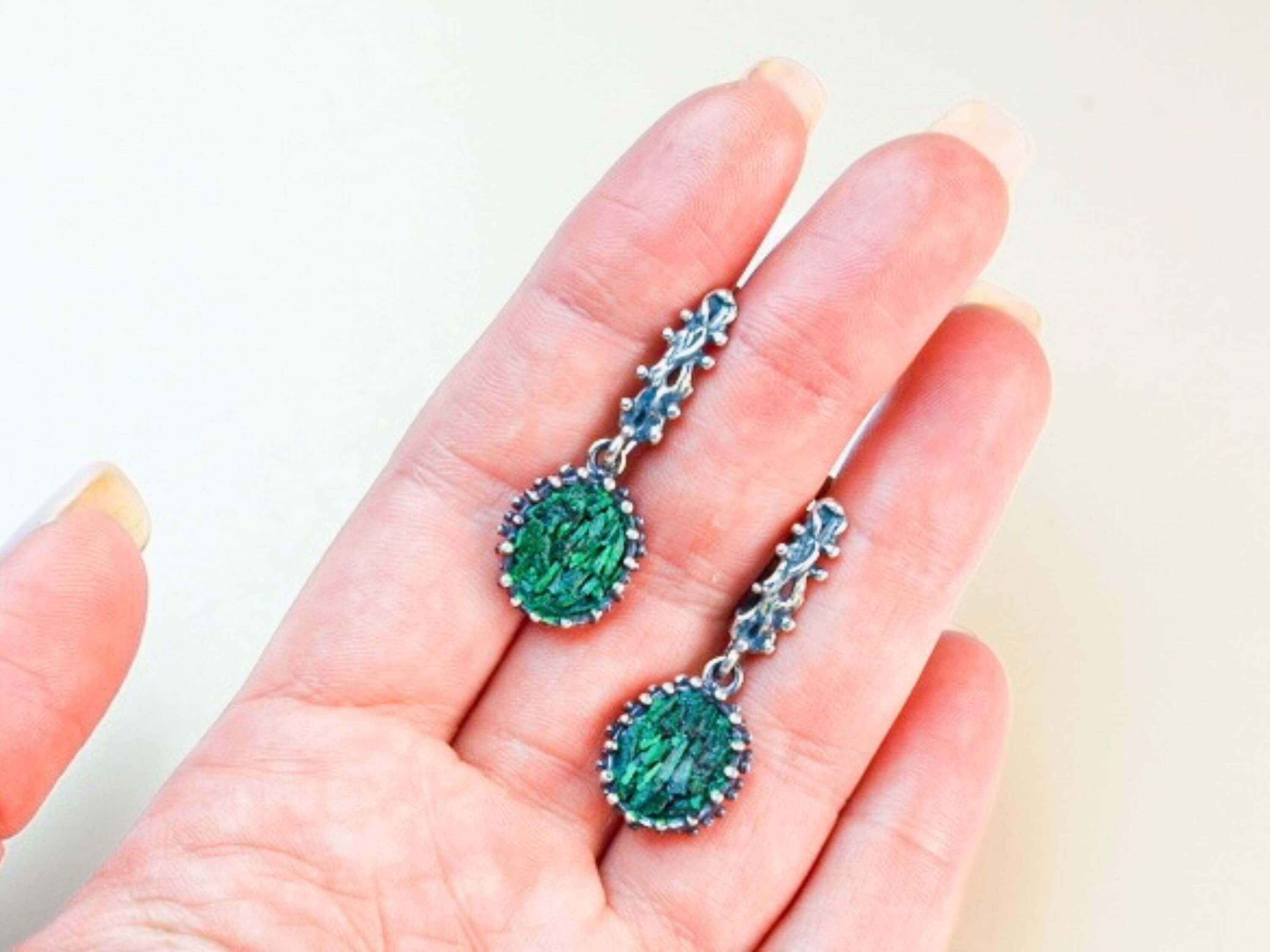 A pair of Akra Gems handcrafted oval Malachite earrings held in a hand to demonstrate the physical scale and artisan craftsmanship