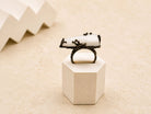 A bold raw white Moonstone statement ring set in a dark oxidized sterling silver frame, displayed on a minimalist geometric stand.